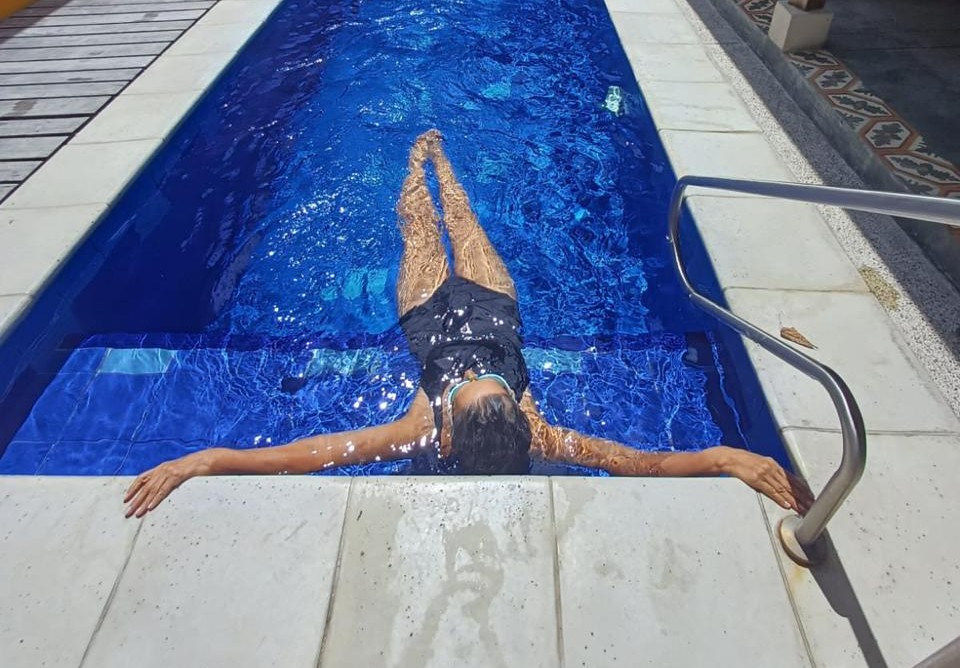 A woman in a black swimsuit relaxes in a blue backyard swimming pool with arms outstretched.
