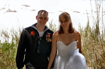 A smiling Navy groom in uniform and bride in a white wedding dress standing on a beach.
