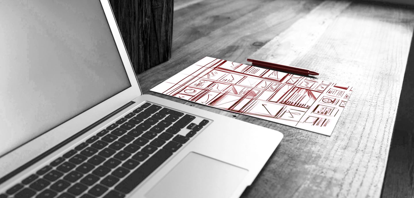 A laptop on a wooden desk next to a sheet of paper with red wireframe designs