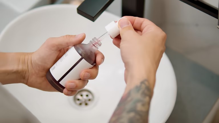 Hands holding a glass dropper bottle with liquid, removing the dropper over a bathroom sink.