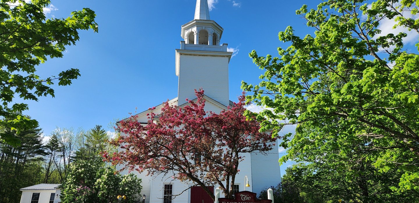 The historic East Winthrop Baptist Church with white siding, a steeple, and a flowering pink tree.