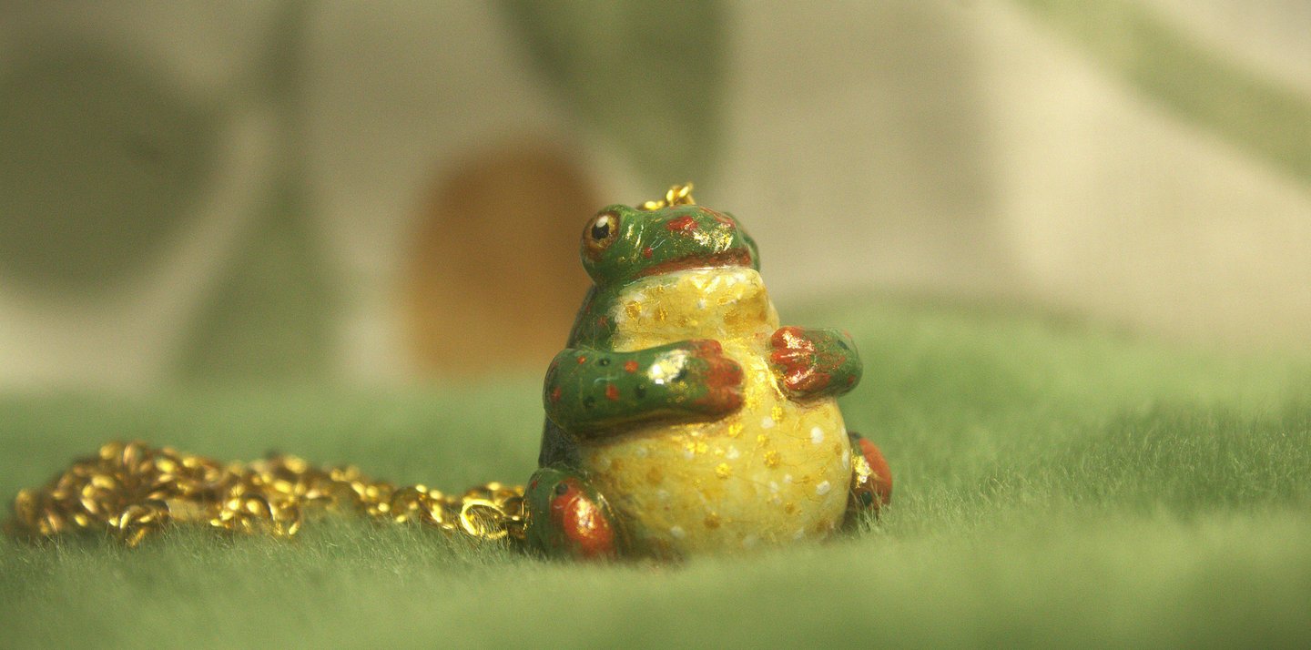 Close-up of a small polymer clay green and gold frog pendant necklace on soft fabric background