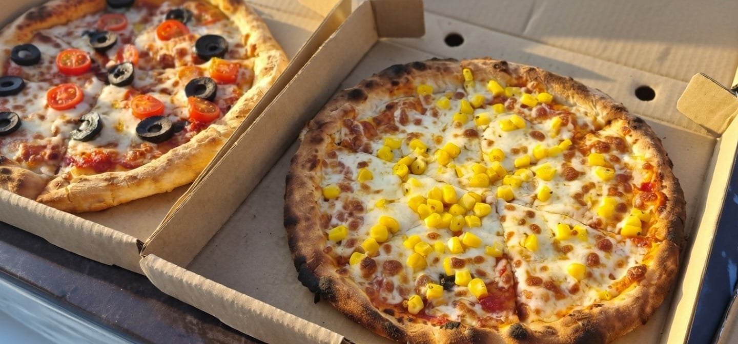 Two takeaway wood-fired pizzas in cardboard boxes with toppings of corn, olives, and cherry tomatoes.