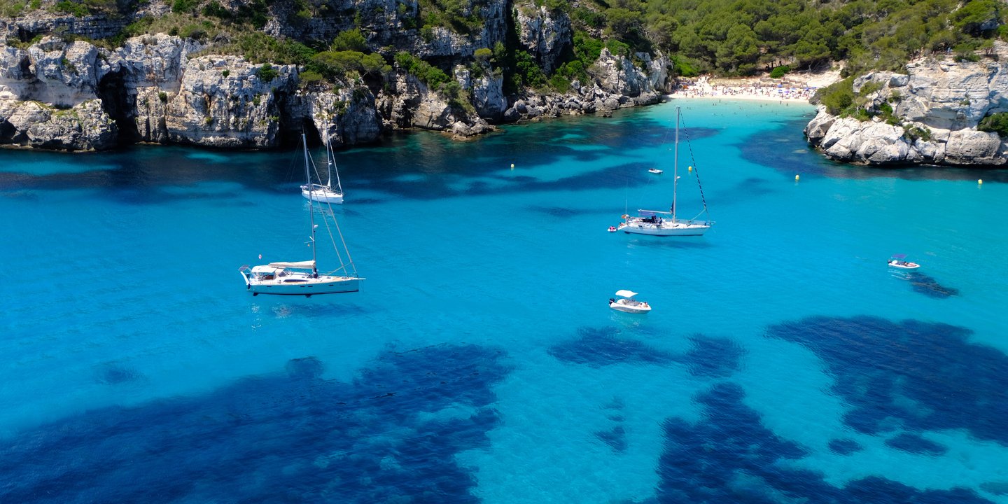 Aerial view of sailboats in the crystal clear turquoise water of Cala Macarella beach in Menorca.