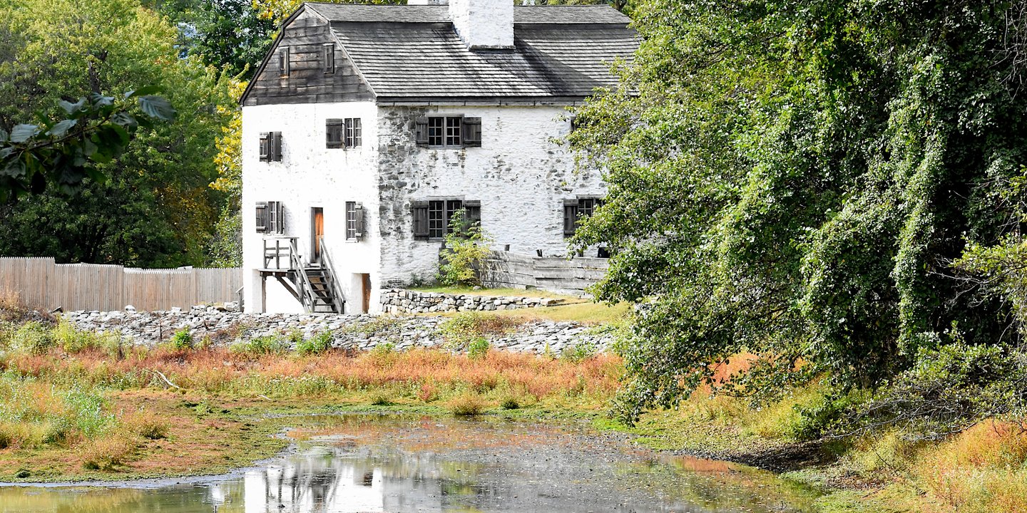 Historic white stone manor house with wooden shutters situated behind a scenic pond and lush green trees.