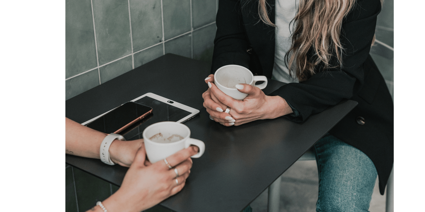 Two women holding coffee mugs while sitting at a cafe table with a smartphone and tablet.