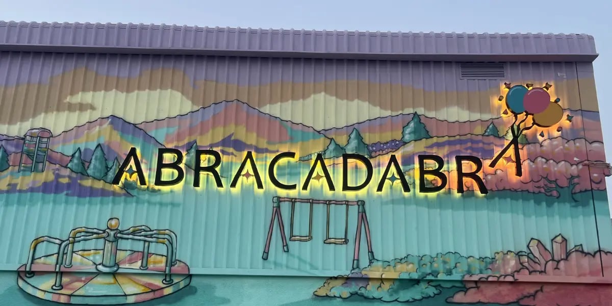 Backlit Abracadabra sign on a colorful playground mural with painted mountains and balloons.