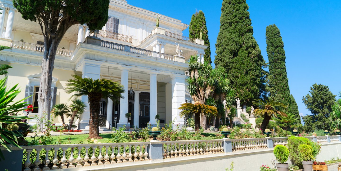 The white Neoclassical facade of Achilleion Palace in Corfu, Greece, surrounded by lush gardens and cypress trees.