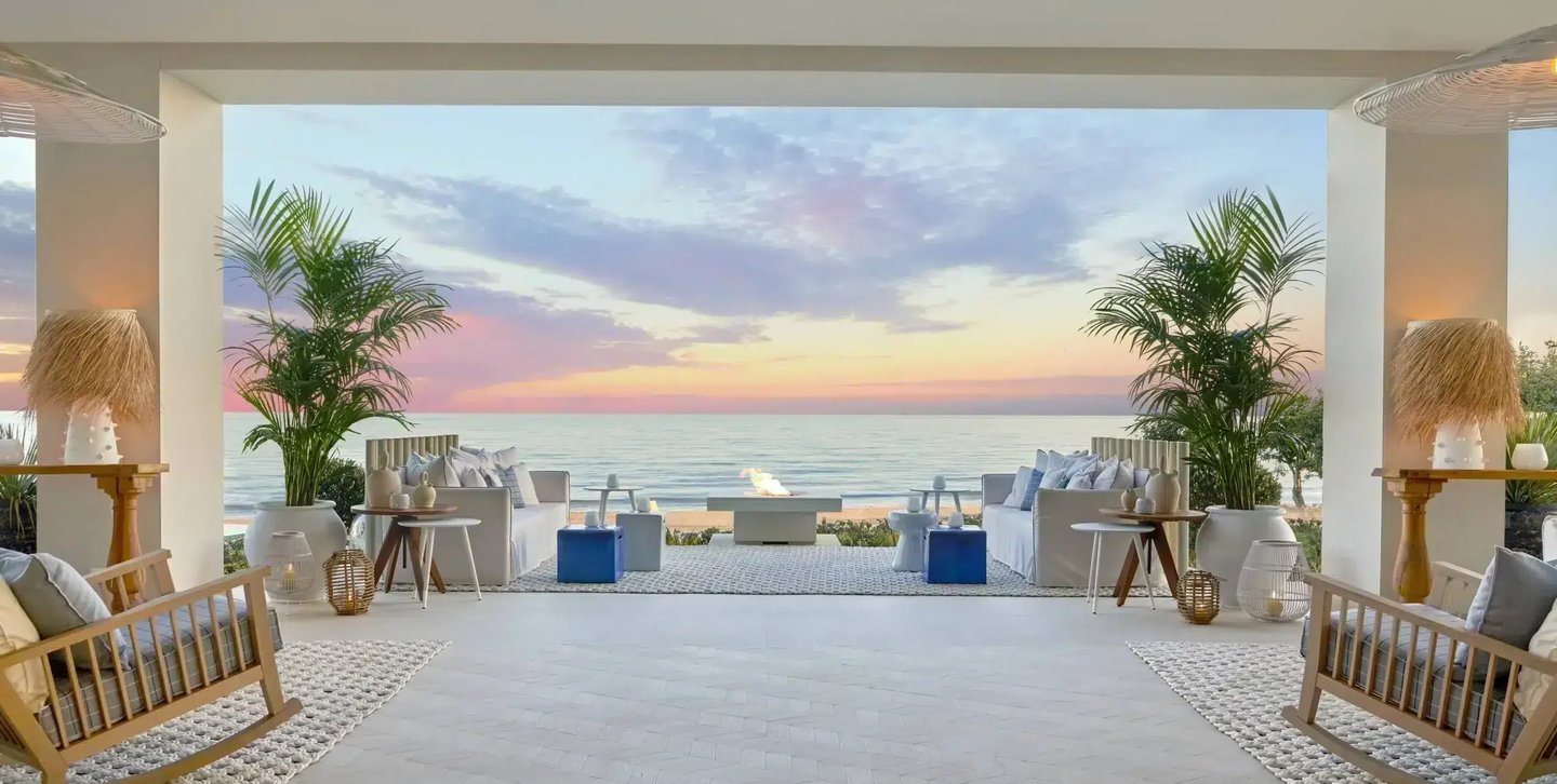 Luxury outdoor lounge with beach views, white sofas, and palm trees during a sunset at a coastal resort.