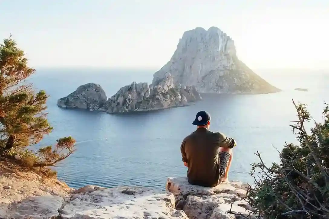 Homme avec 1 casquette visière en AR assis sur 1 rocher face à 1 gros promontoir rocheux dans la mer