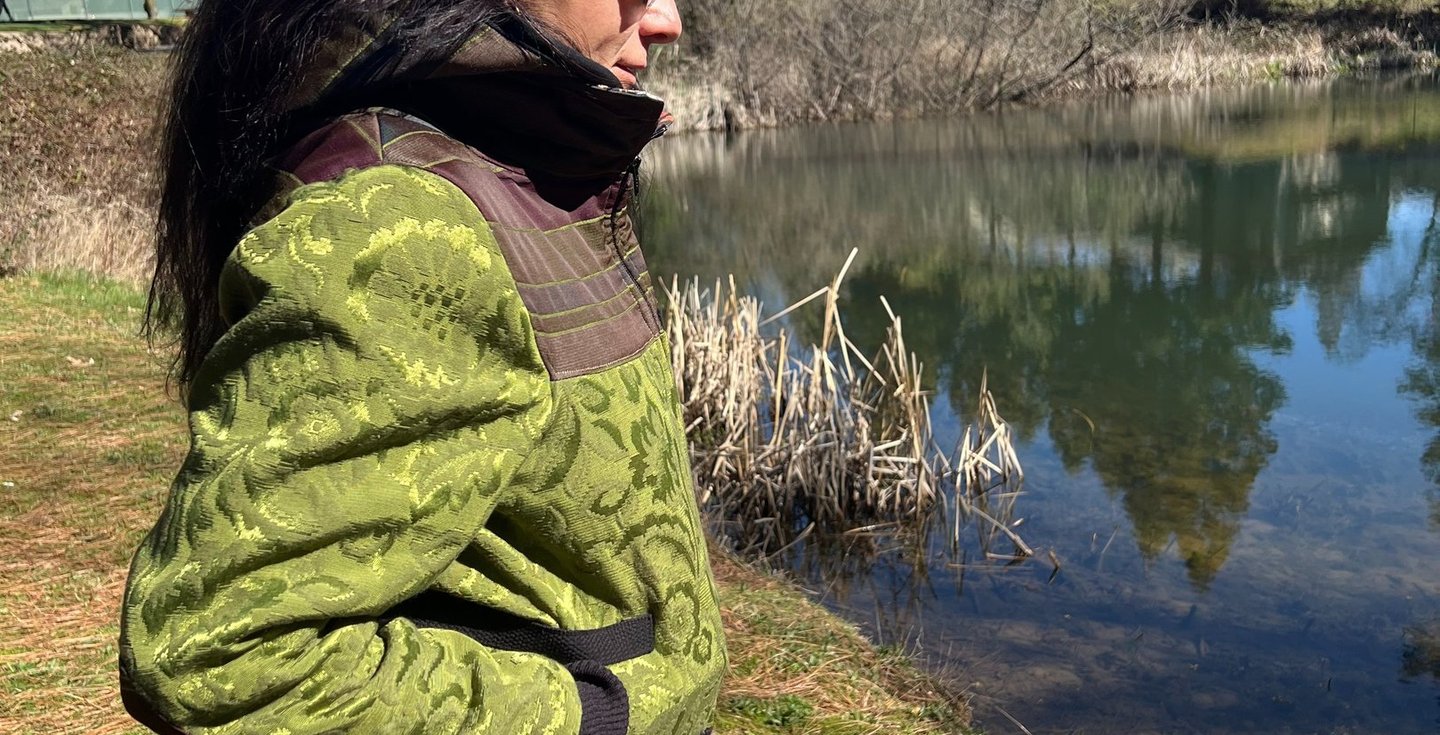 a woman in a green jacket and jeans standing in front of a pond