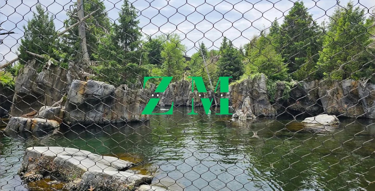A naturalistic zoo habitat with a water pond and rocky cliffs viewed through a wire mesh fence.