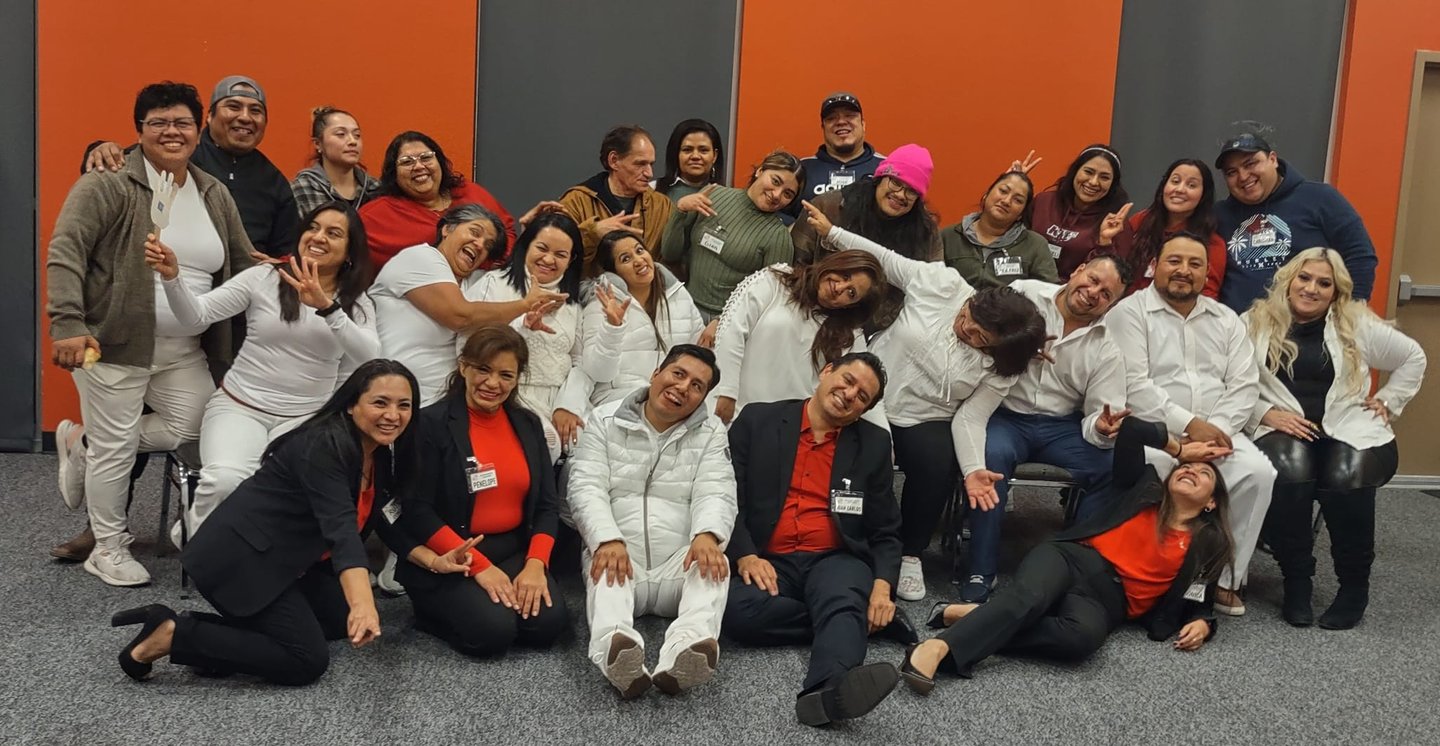 A diverse group of cheerful coworkers posing for a team photo in an office with an orange and grey wall.