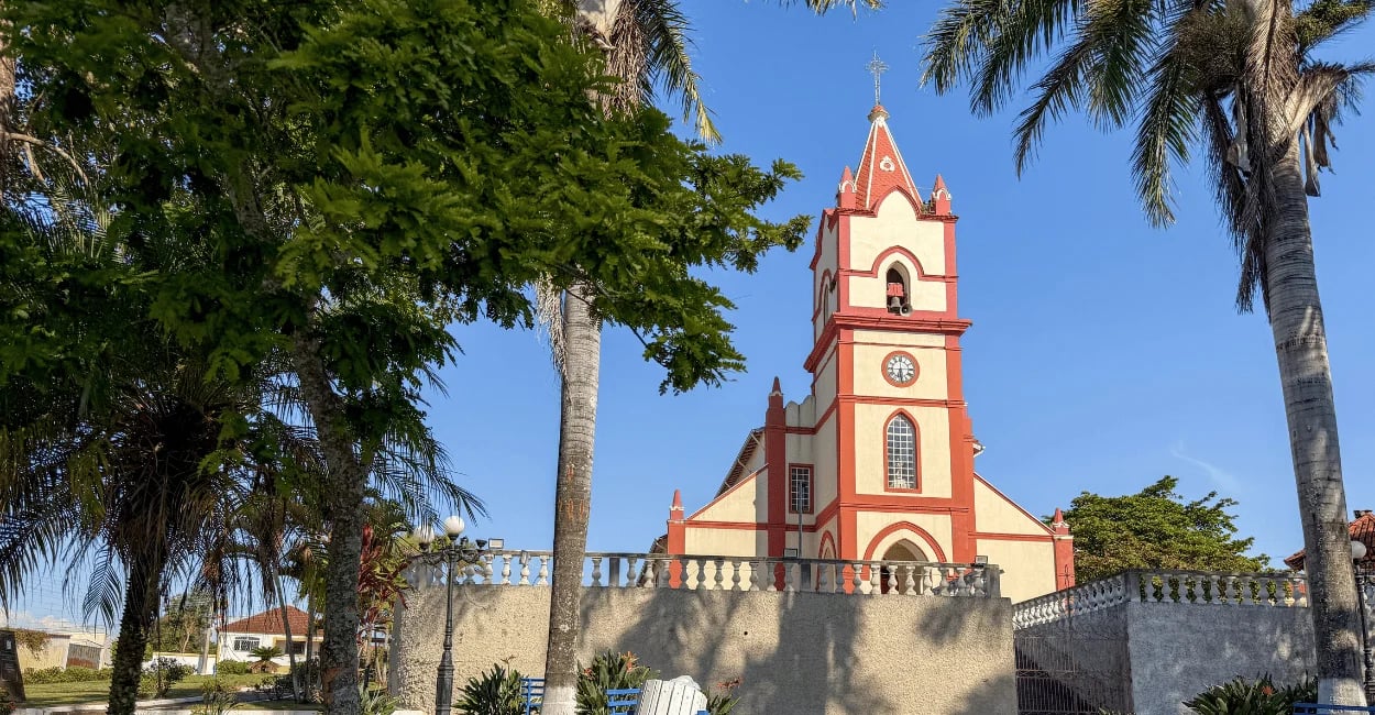Igreja Matriz de Ibituruna cercada por palmeiras, com torre central, relógio e fachada vermelha e br