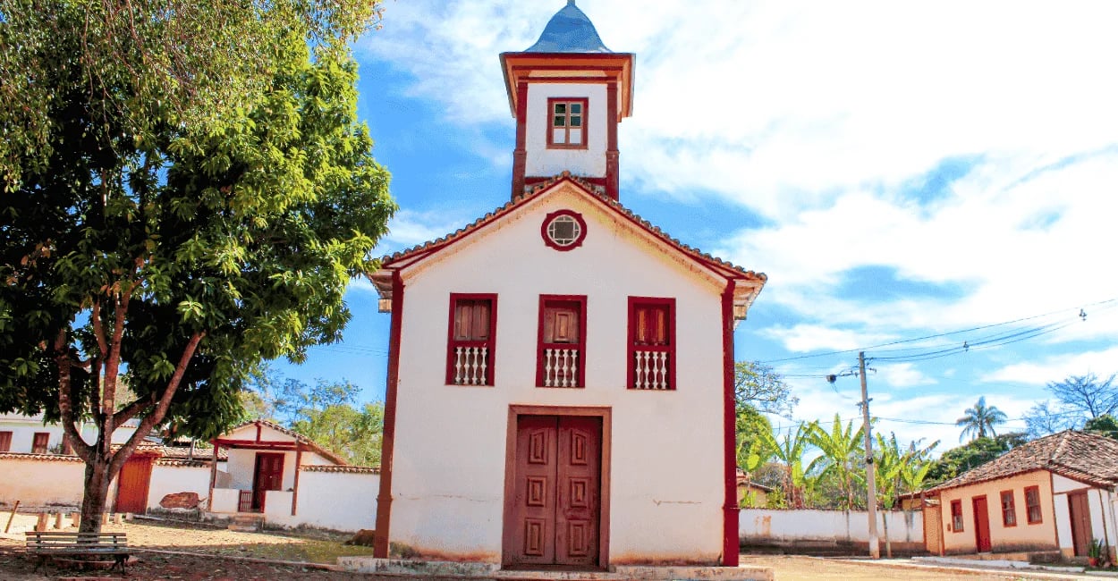 Pequena capela histórica de fachada branca e portas de madeira em uma praça arborizada no distrito
