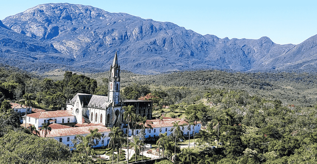 Santuário do Caraça com sua torre imponente, montanhas e vegetação ao fundo, com céu aberto