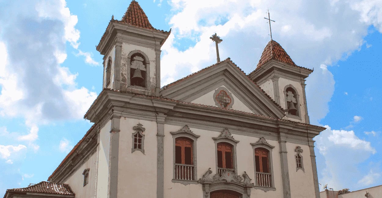 Igreja Matriz de Santa Bárbara MG com duas torres, fachada clara e céu azul com nuvens ao fundo