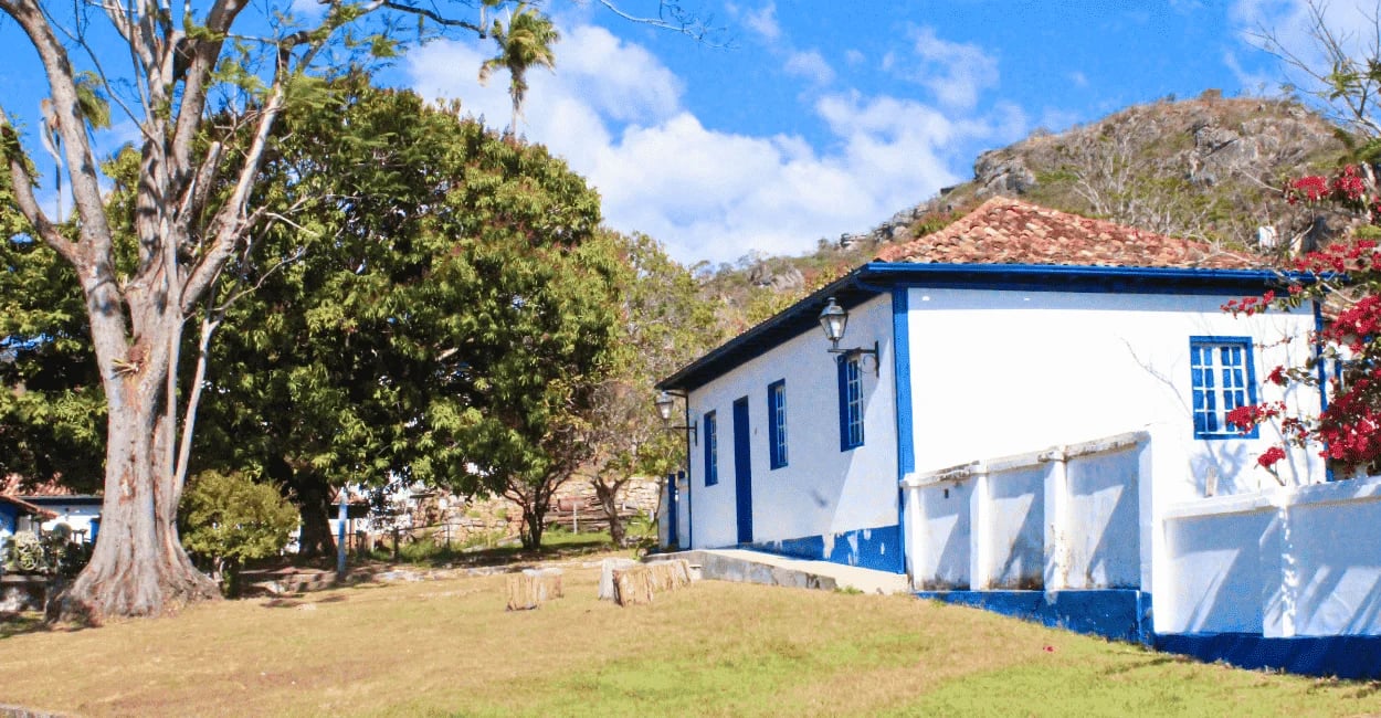 Casa histórica na Vila do Biribiri, com fachada branca e azul, jardim amplo, árvores e serra ao fund