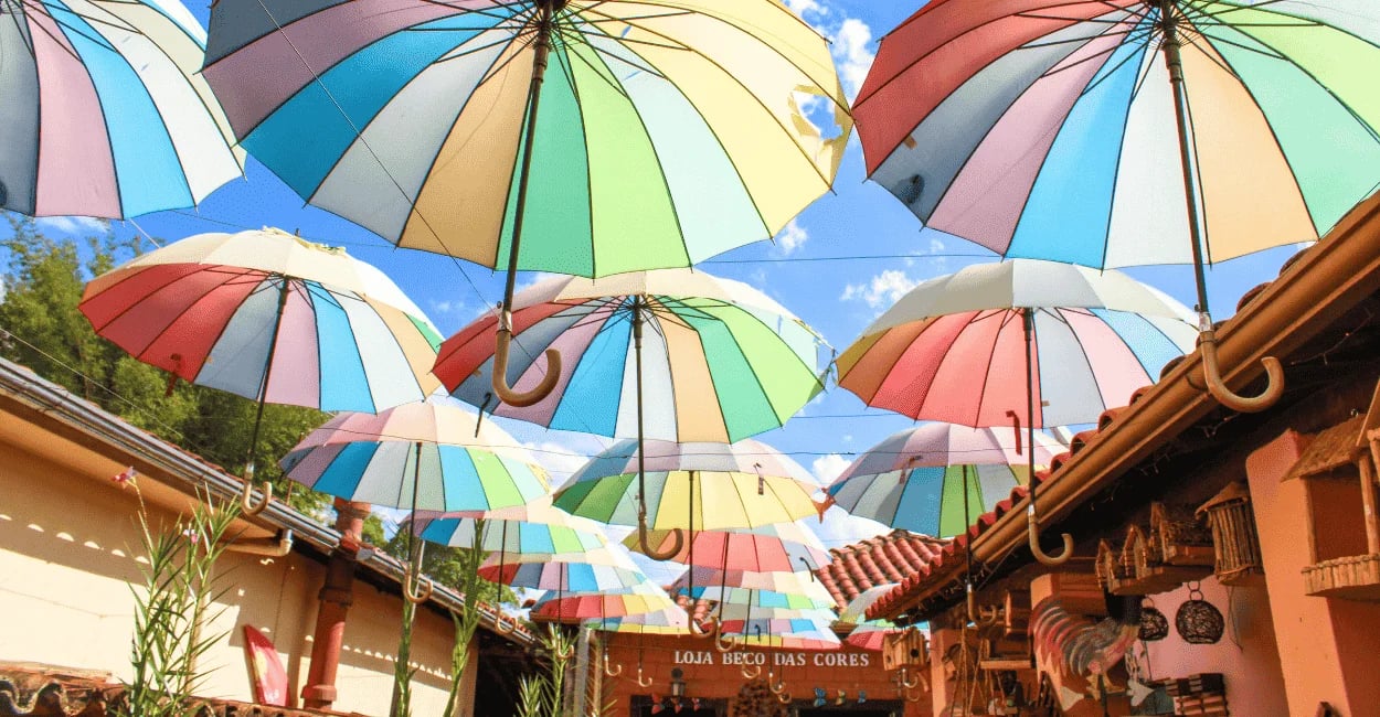 Guarda-chuvas coloridos suspensos sobre rua de lojas em Bichinho, MG, criando um corredor