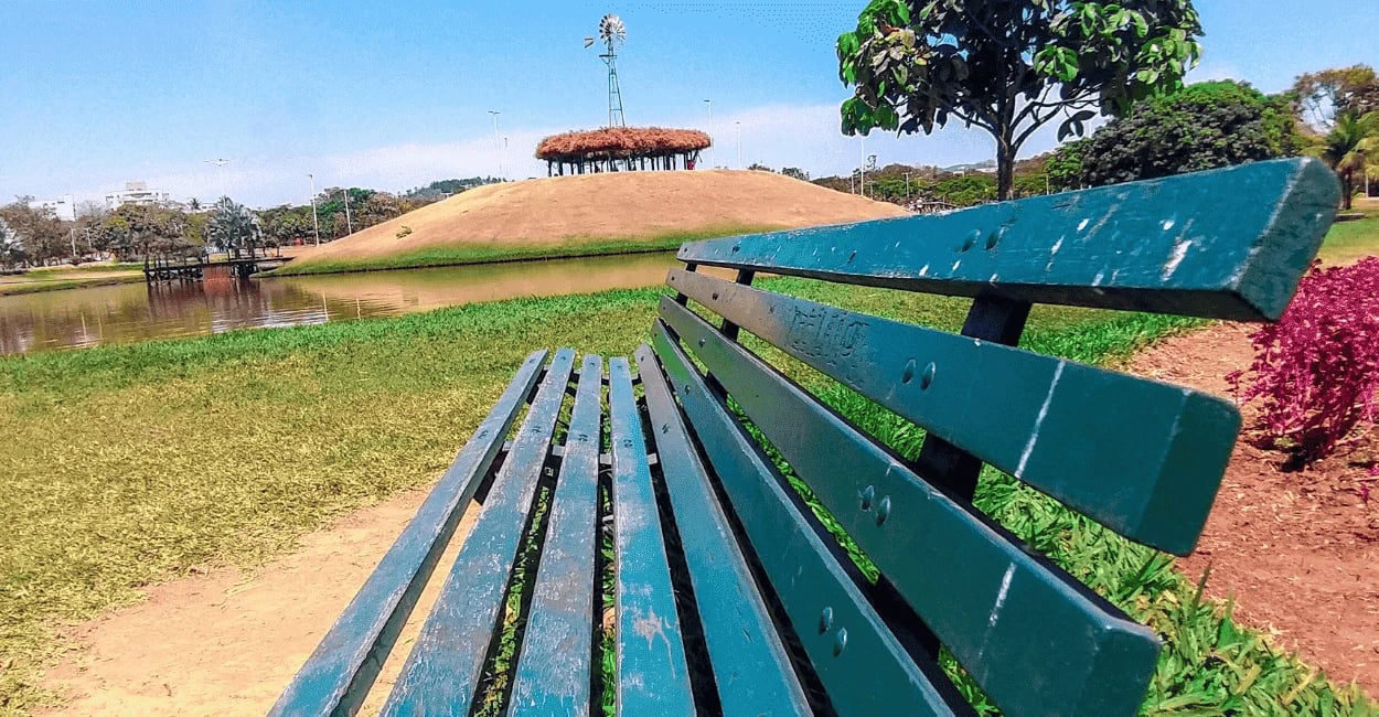 Banco de cor verde, água, ilha com moinho de vinho no alto e céu azul, no Parque Ipanema em Ipatinga