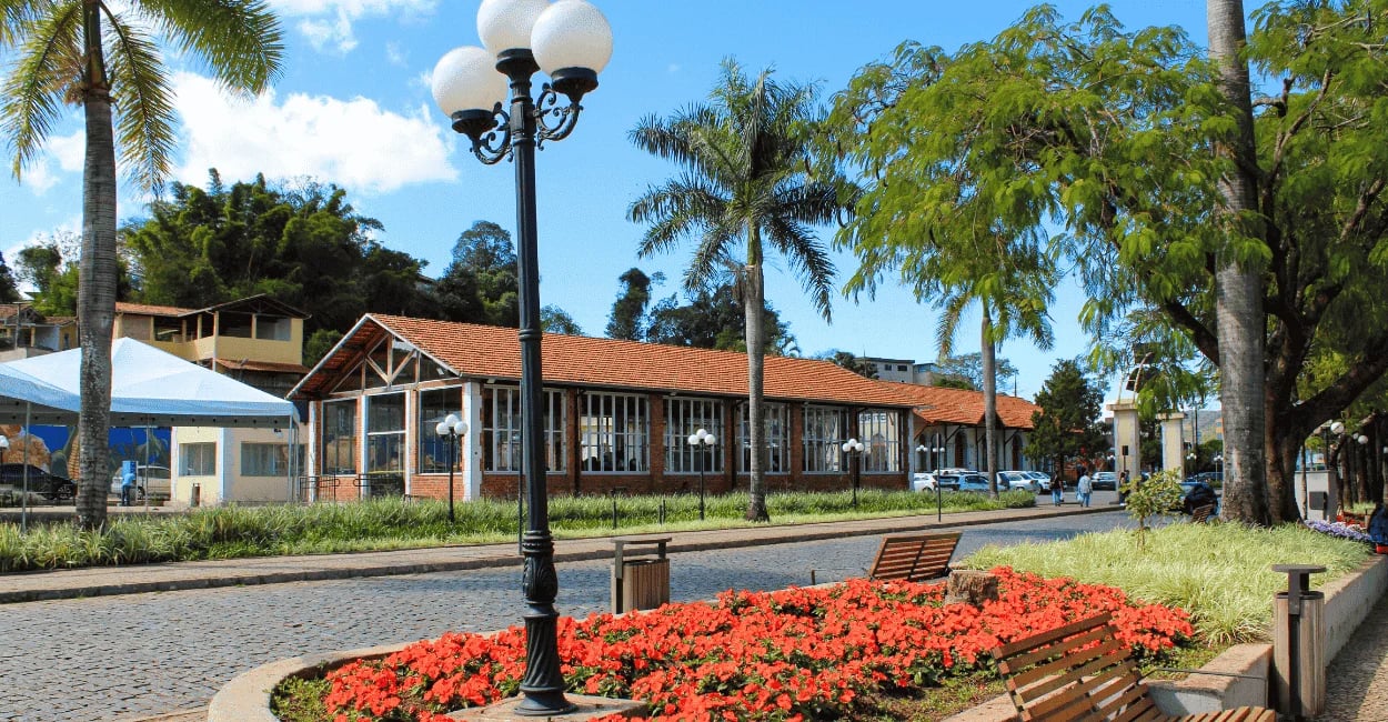 Praça arborizada em Itabirito, MG, com canteiro de flores vermelhas, bancos e museu ferroviário