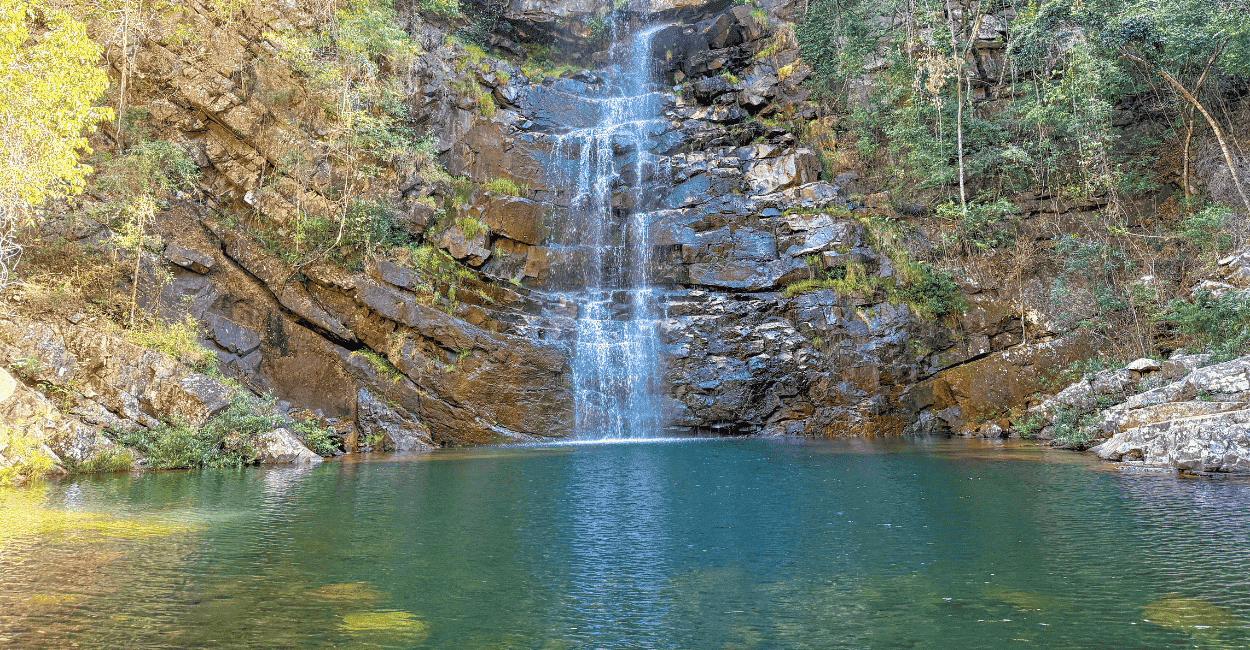 Uma queda d'água de uma cachoeira com um poço de água verde claro, em um paredão e vegetação