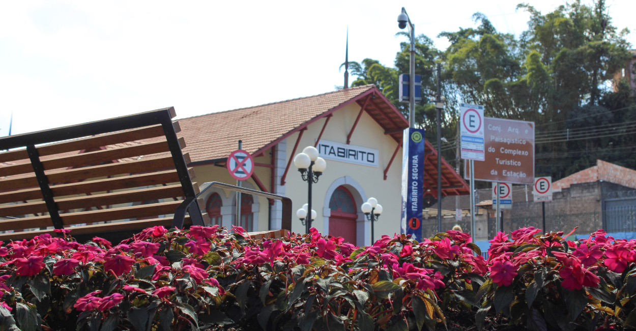 Praça Central de Itabirito, com flores e um banco, com a estação ferroviaria histórica