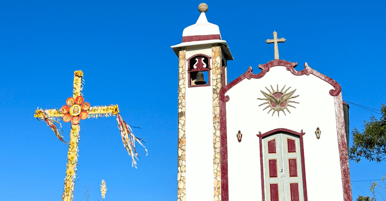 Cruz e uma capela de cor branca e vinho, com céu azul no Morro Redondo de Ipoema MG