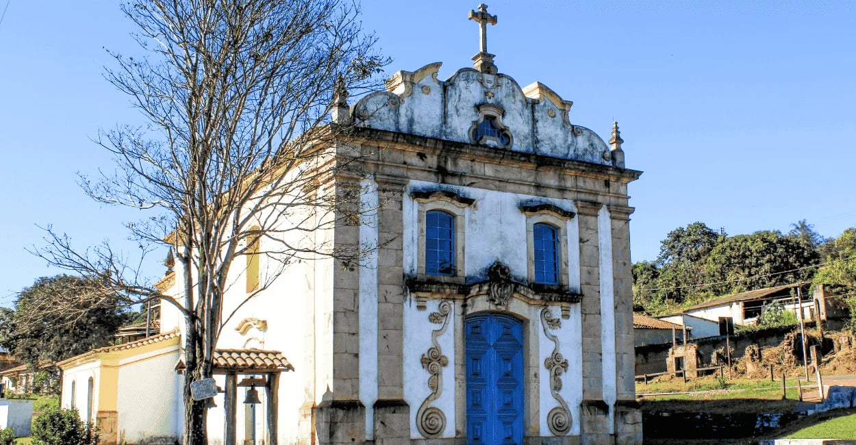 Pequena igreja histórica branca e azul ao lado de uma árvore seca em Lobo Leite, MG