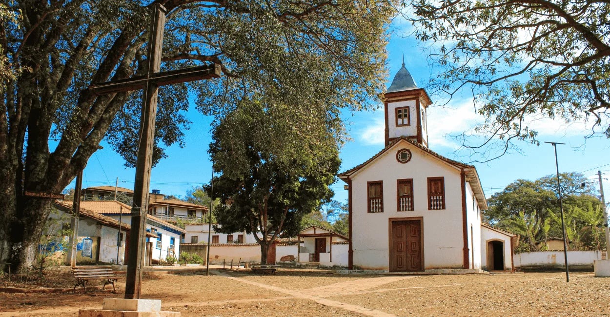 Capela com cores brancas e vermelhas, cruz de madeira a frente e arvores em uma praça em MG