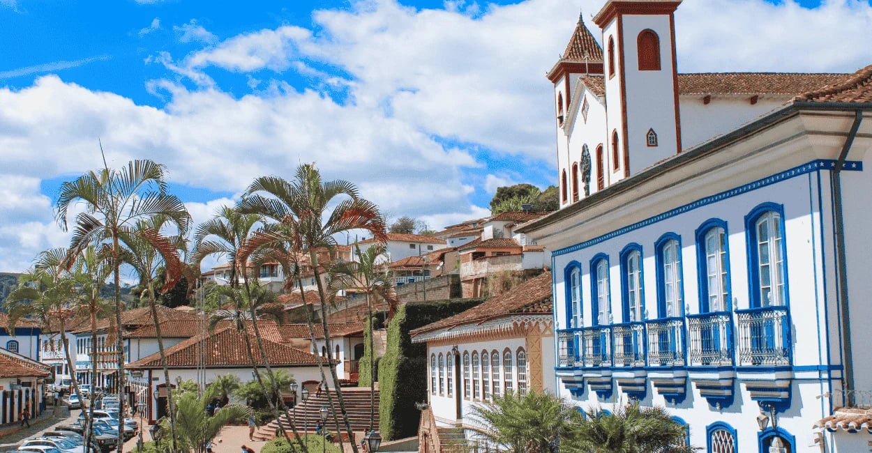 Casario do Serro, em MG, com fachadas azuis, palmeiras e igreja histórica no alto, sob céu azul