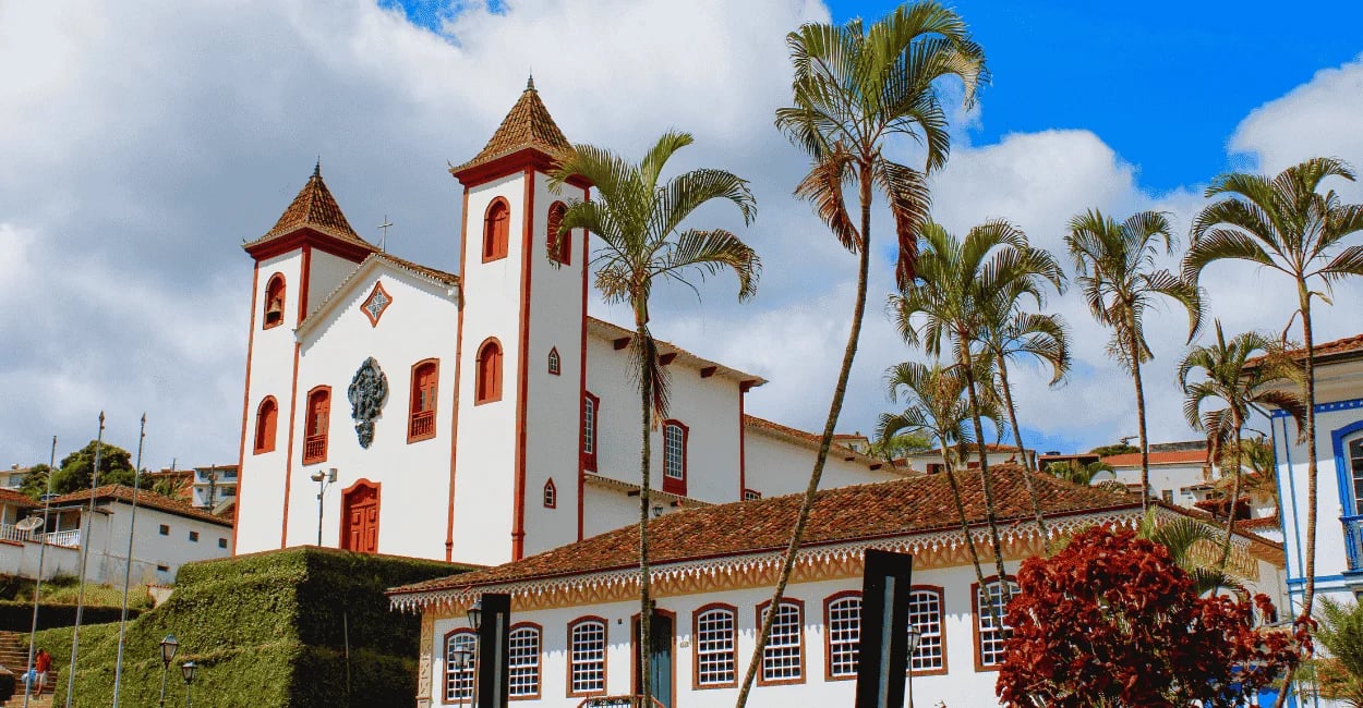 Igreja histórica em Serro com fachada branca, detalhes vermelhos, palmeiras altas e céu nublado