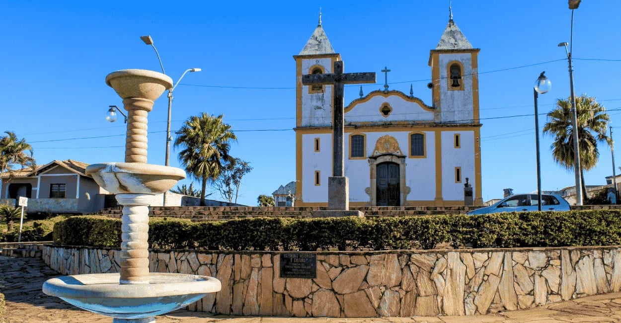 Fonte na praça de Queluzito com igreja matriz amarela e branca ao fundo em Minas Gerais