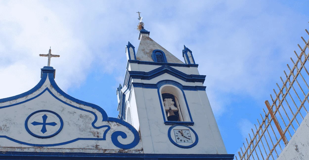 Cruz e torre da Igreja Matriz de Raposos, em Minas Gerais, com fachada branca e detalhes azuis