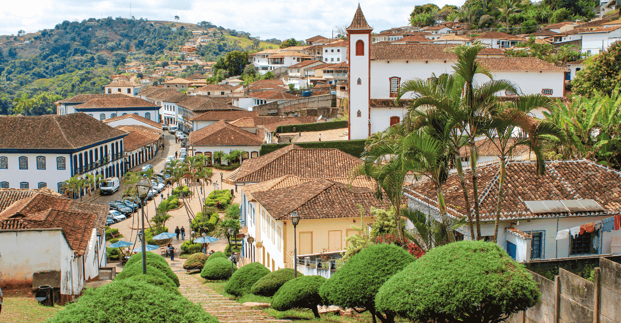 Uma escadaria descendo com pequenas árvores ao lado, casas historicas ao fundo e uma Igreja do canto
