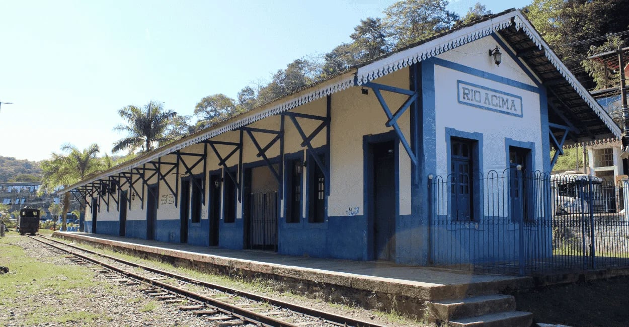 Antiga estação ferroviária de Rio Acima com fachada branca e azul, plataforma comprida e trilhos