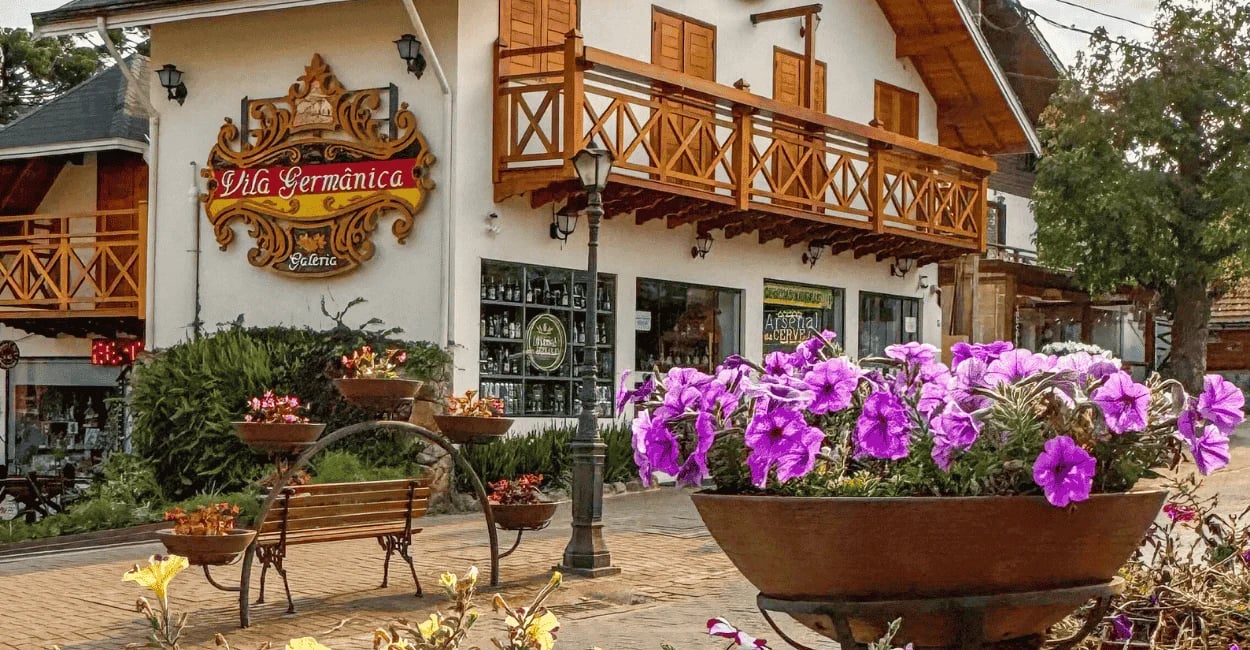 Fachada de edifício em estilo germânico com floreiras de petúnias roxas na calçada em Monte Verde