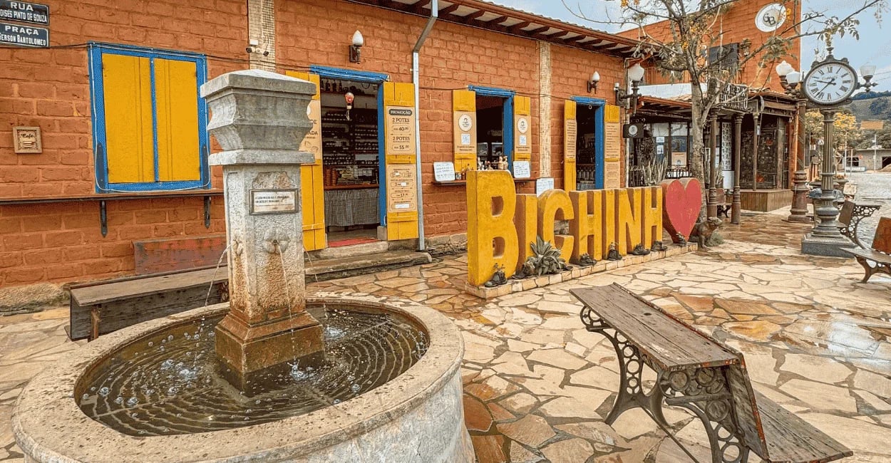 Praça em Bichinho MG com chafariz de pedra, letreiro turístico, casarões coloridos e relógio de rua
