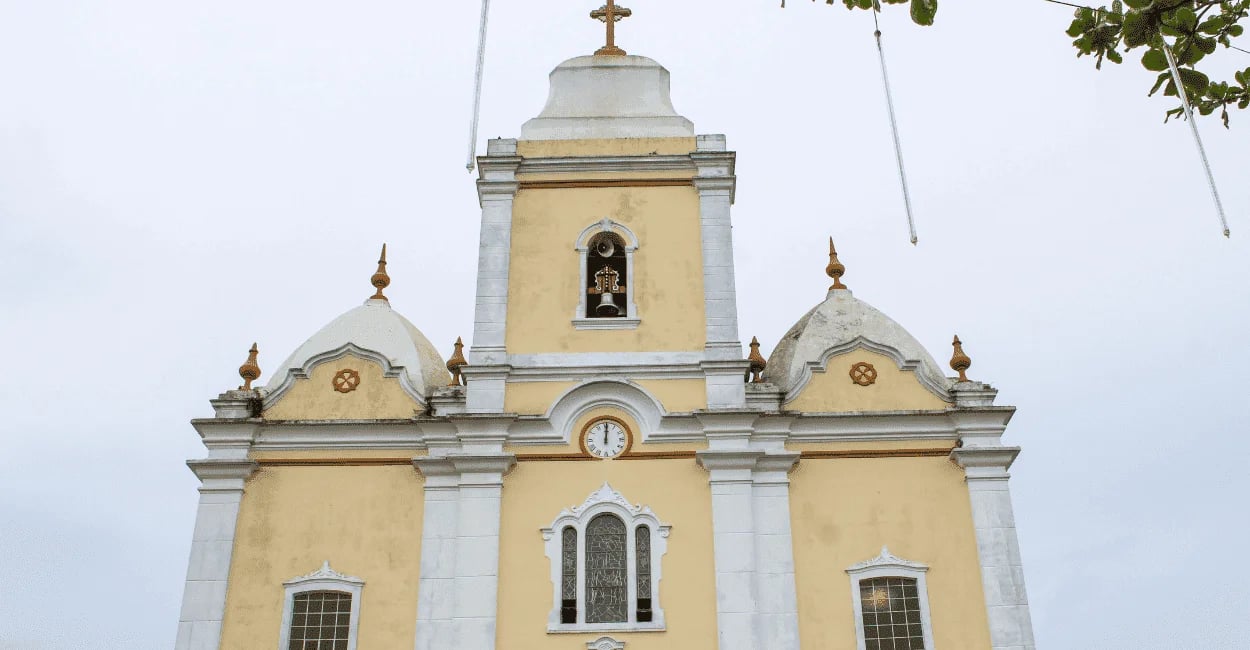 Foto frontal da Igreja Matriz de Resende Costa, em cores branca e amarela, com um sino na torre