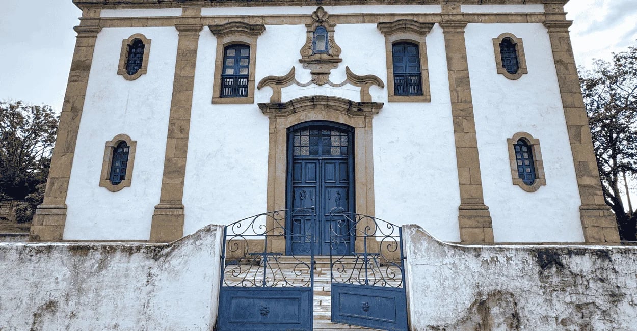 Fachada da Igreja Matriz de Glaura MG com porta e portão azul, detalhes em pedra e muro antigo