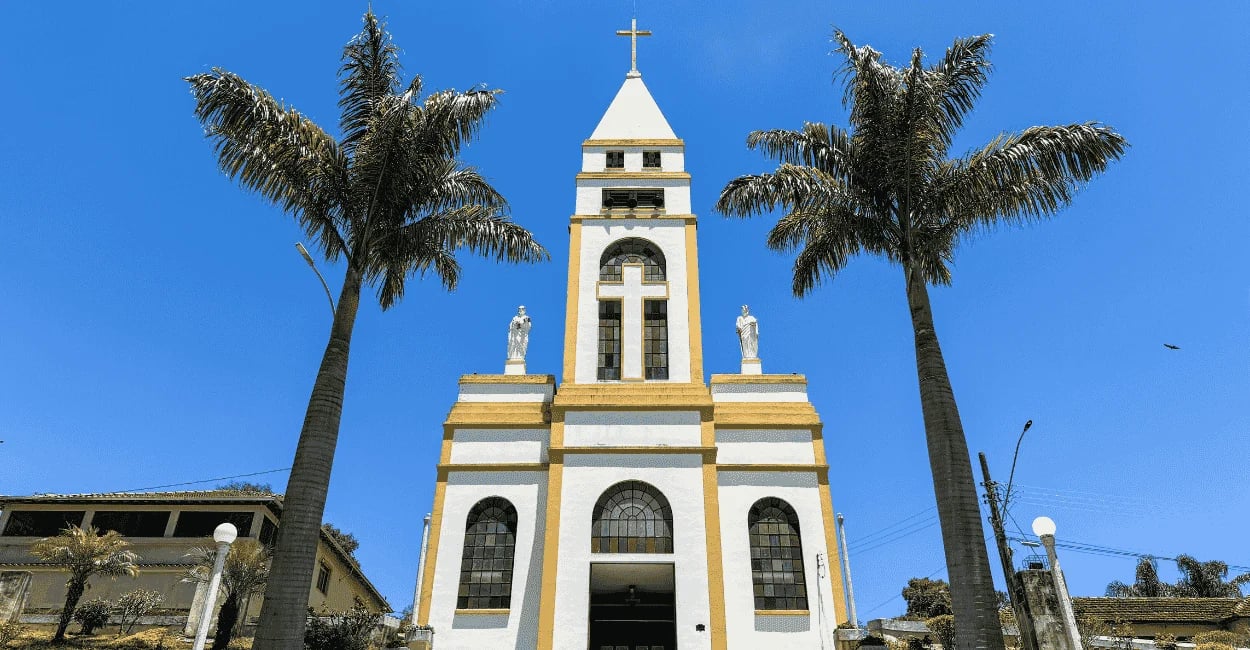 Fachada da igreja branca e amarela ladeada por duas palmeiras altas em Ressaquinha, MG
