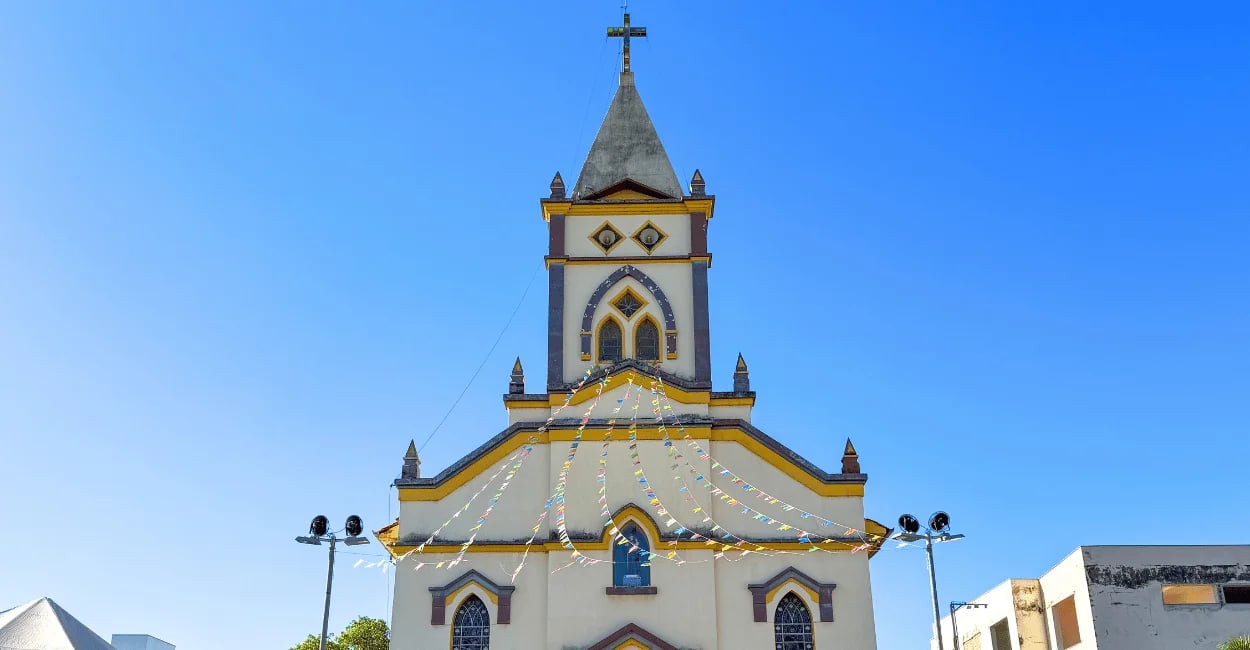 Fachada da Igreja Matriz de Igarapé com torre central, cruz e bandeirinhas coloridas sob céu azul