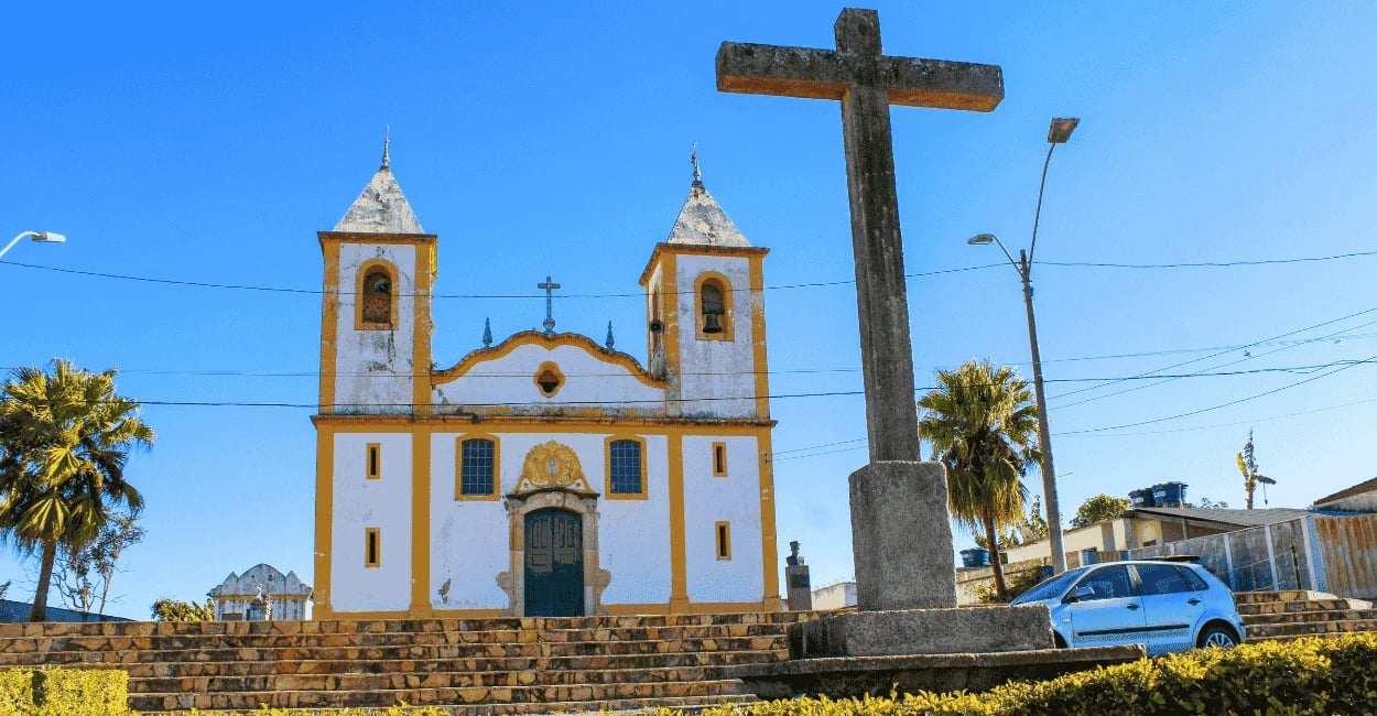 Fachada da Igreja Matriz branca e amarela com escadaria e cruz de pedra. Queluzito, MG