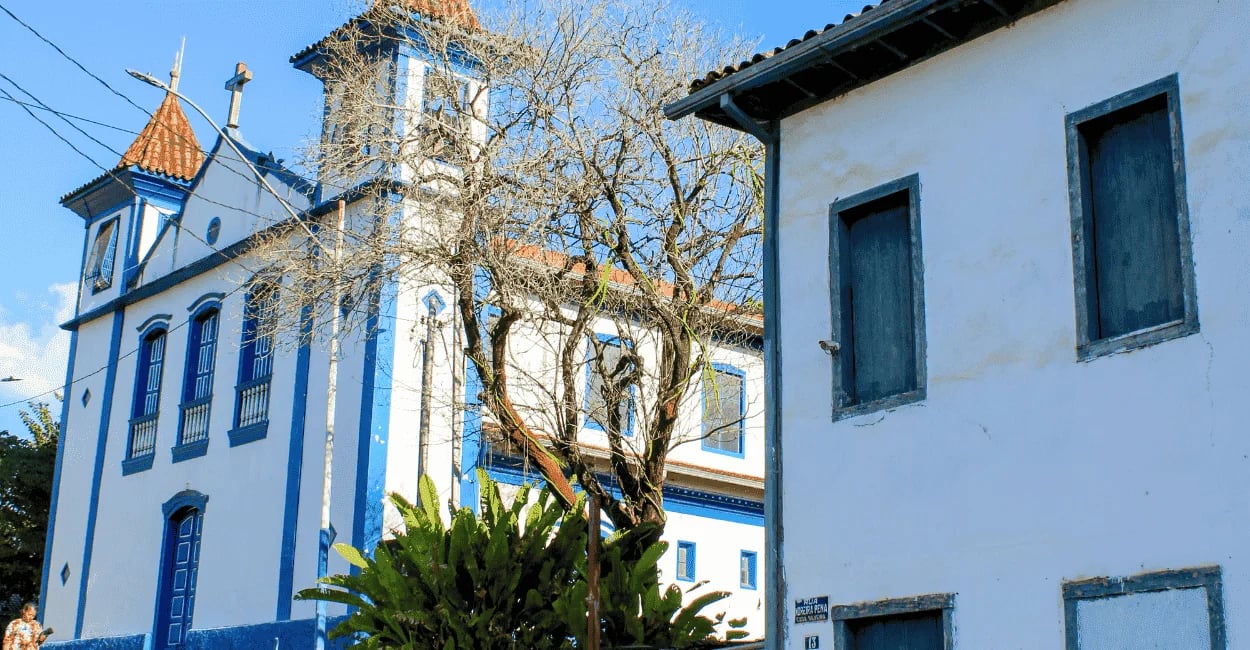 Igreja histórica em Ipoema, com fachada branca e azul, torre sineira, árvores sem folhas e casarão