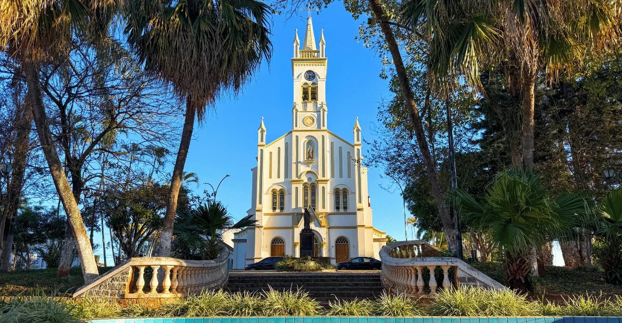 Igreja Matriz de Carmópolis de Minas vista de frente entre árvores e jardins da praça central