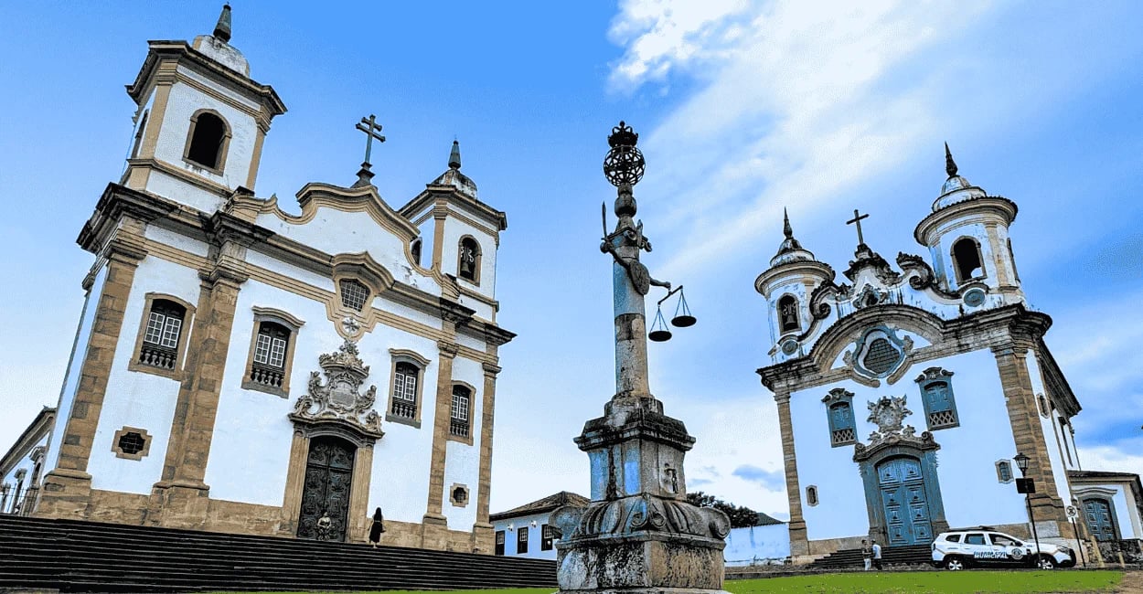 Igrejas históricas na Praça Minas Gerais em Mariana, com pelourinho ao centro e céu azul ao fundo