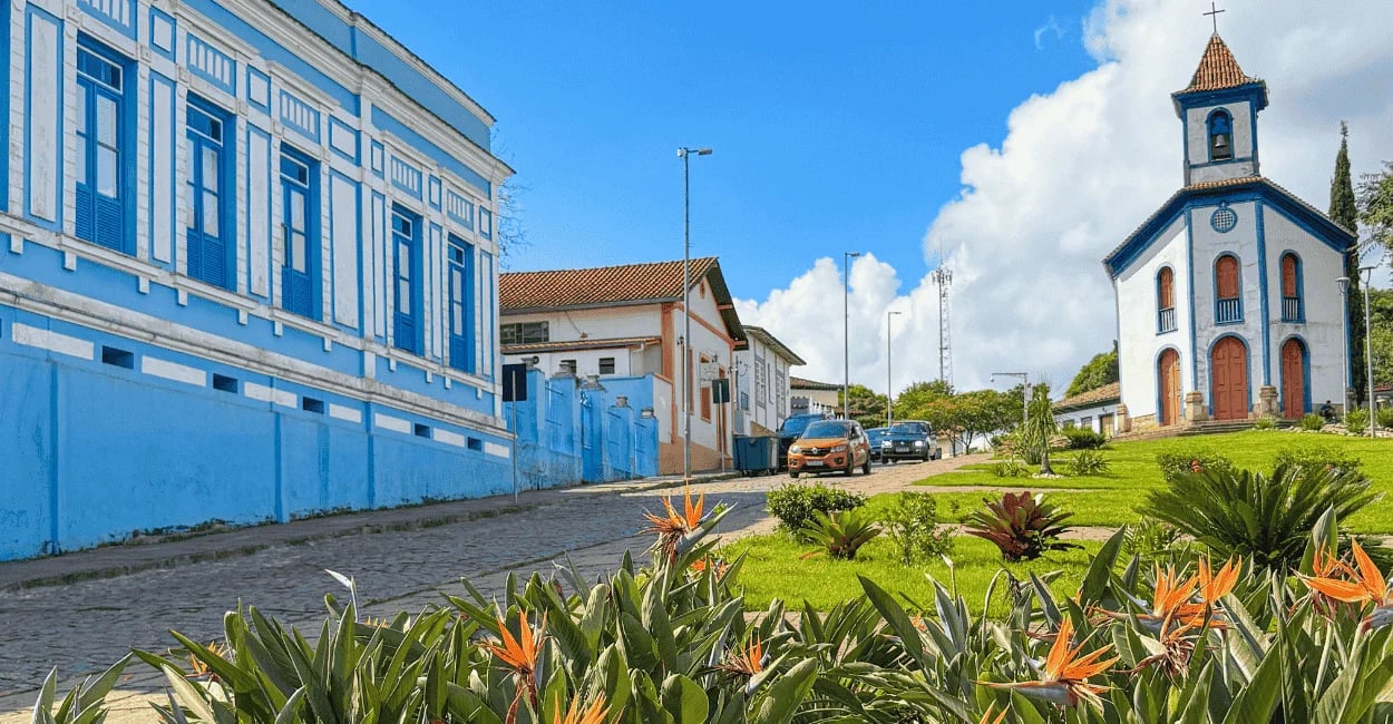 Rua de Santa Bárbara com casarão azul, igreja em destaque e jardim florido em primeiro plano
