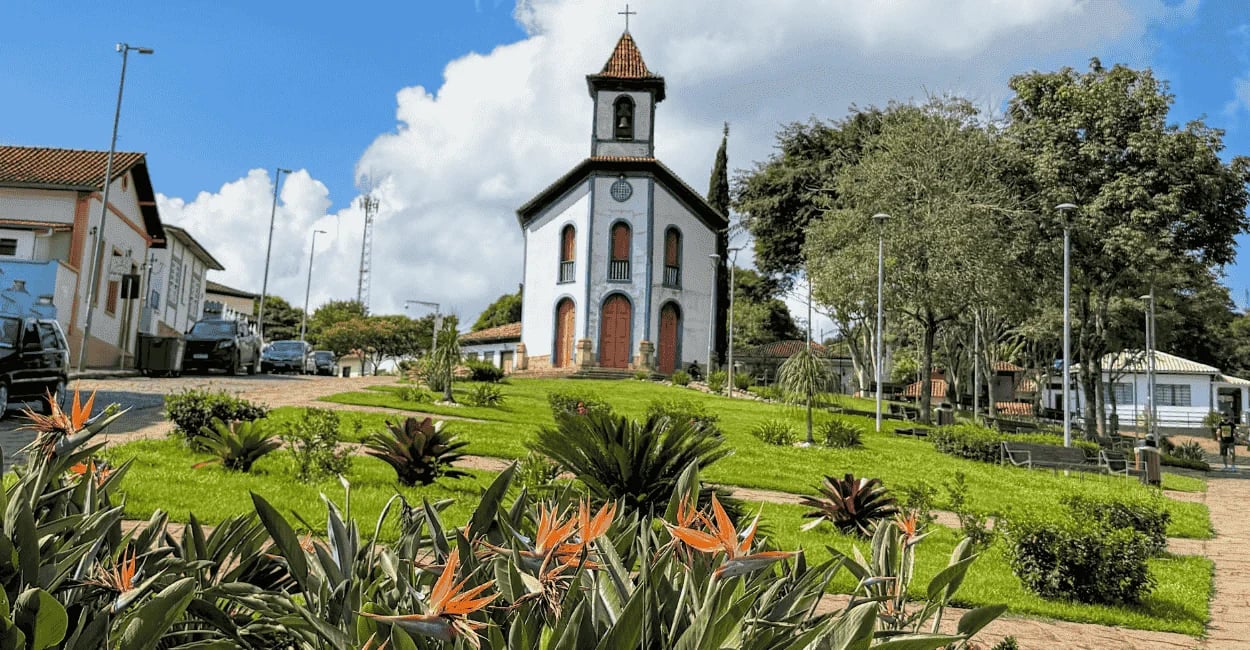 Igreja em Santa Bárbara vista da praça, com flores em primeiro plano, gramado e céu azul com nuvens