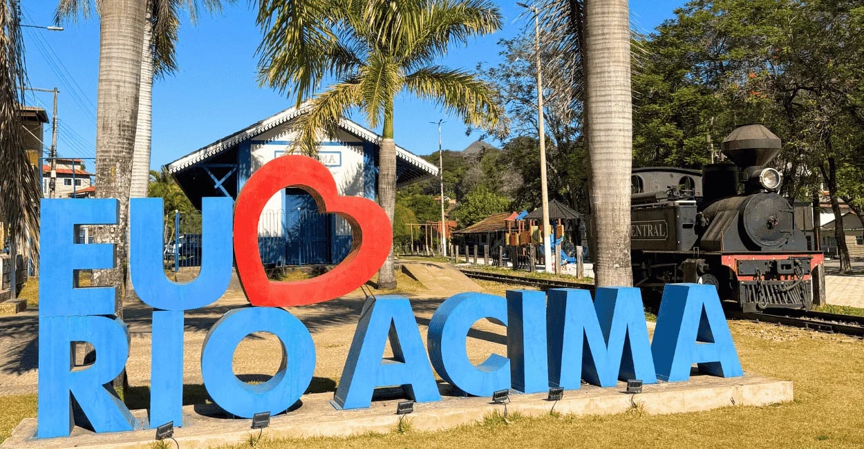 Letreiro de Rio Acima em frente à antiga estação ferroviária, com locomotiva histórica e palmeiras
