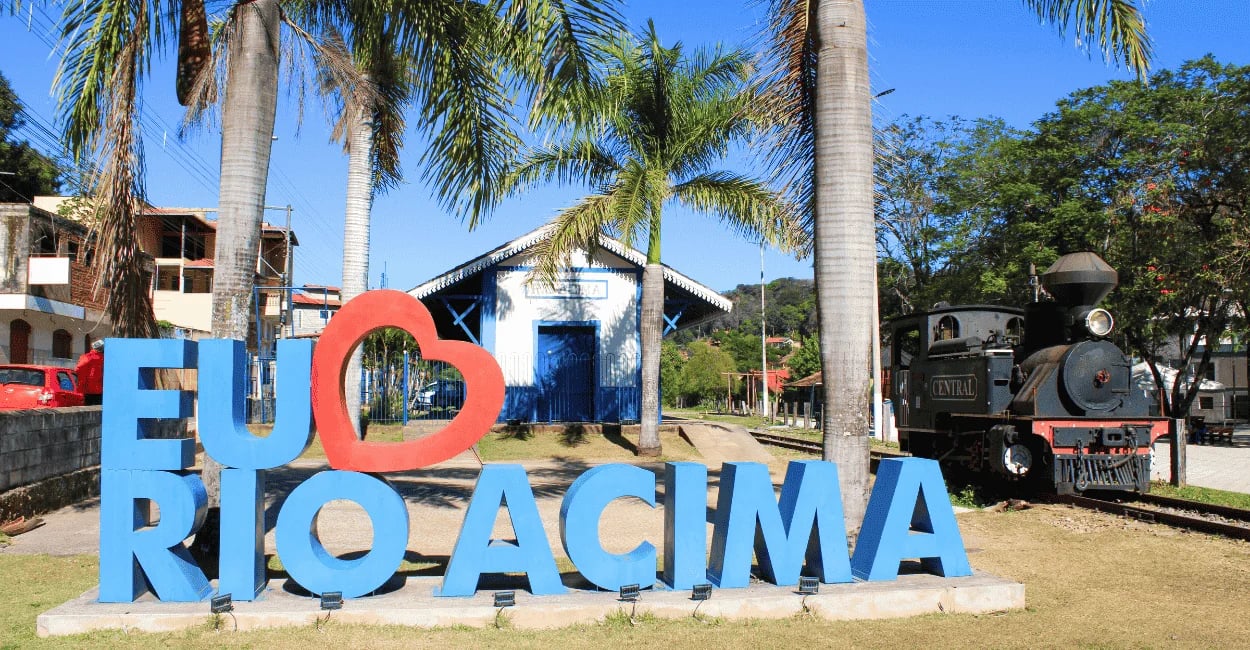Locomotiva a vapor antiga estacionada ao lado de letreiro turístico de Rio Acima e serra ao fundo